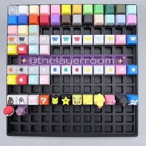 Peut inclure: Une collection de keycaps color&eacute;es dispos&eacute;es sur une grille noire. Les keycaps pr&eacute;sentent diverses couleurs et motifs, dont la lettre "W", le chiffre "8" et des personnages de dessins anim&eacute;s. Le texte "@thelayerroom" est &eacute;galement visible.