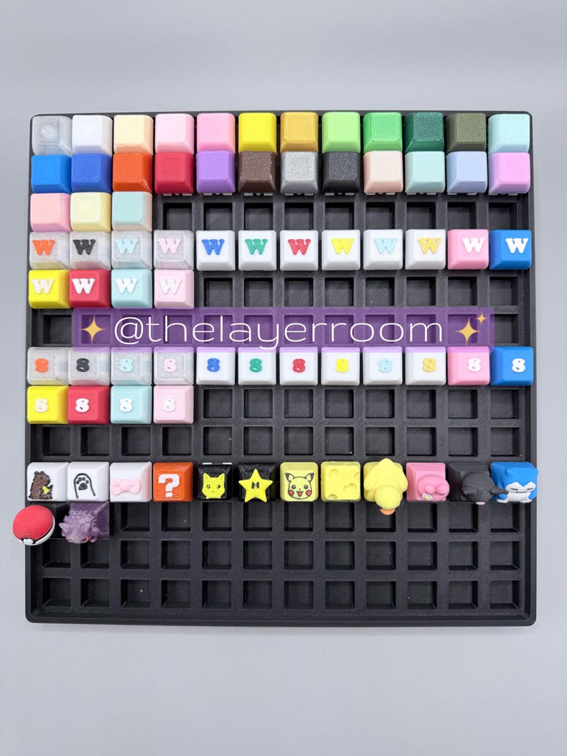 Peut inclure: Une collection de keycaps color&eacute;es dispos&eacute;es sur une grille noire. Les keycaps pr&eacute;sentent diverses couleurs et motifs, dont la lettre "W", le chiffre "8" et des personnages de dessins anim&eacute;s. Le texte "@thelayerroom" est &eacute;galement visible.