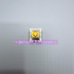 Peut inclure: Un interrupteur de clavier m&eacute;canique unique avec une tige jaune en forme de croix et un bo&icirc;tier transparent. L'interrupteur a une croix dor&eacute;e au centre. Le texte "@thelayerroom" est sur un autocollant violet en dessous.