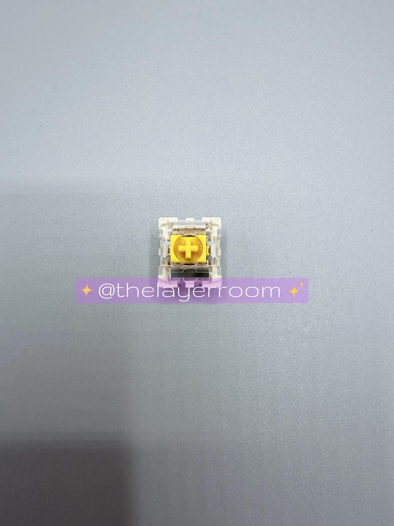 Peut inclure: Un interrupteur de clavier m&eacute;canique unique avec une tige jaune en forme de croix et un bo&icirc;tier transparent. L'interrupteur a une croix dor&eacute;e au centre. Le texte "@thelayerroom" est sur un autocollant violet en dessous.