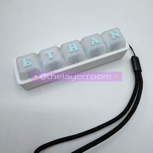 Peut inclure: Un ensemble de touches de clavier personnalis&eacute;es &eacute;pelant "ETHAN" en lettres bleu clair sur des touches transparentes. Les touches sont dans un plateau blanc avec une lani&egrave;re noire attach&eacute;e. Le texte "@thelayerroom" est &eacute;galement visible.