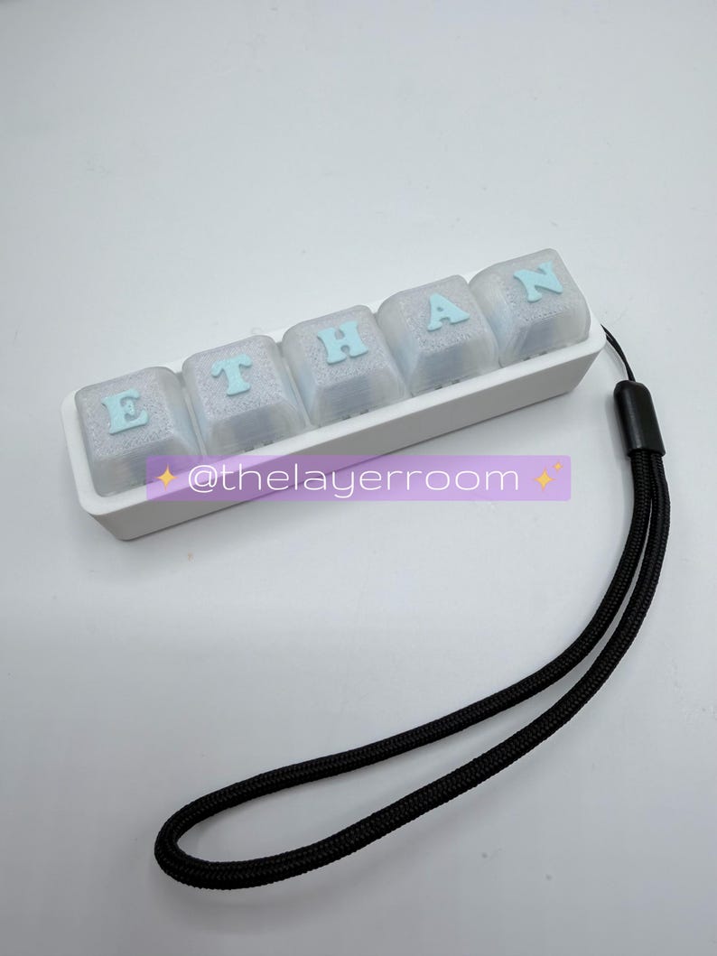 Peut inclure: Un ensemble de touches de clavier personnalis&eacute;es &eacute;pelant "ETHAN" en lettres bleu clair sur des touches transparentes. Les touches sont dans un plateau blanc avec une lani&egrave;re noire attach&eacute;e. Le texte "@thelayerroom" est &eacute;galement visible.