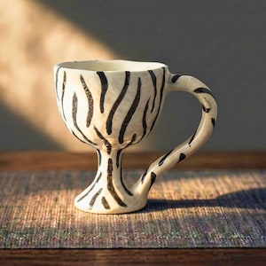 Puede incluir: Una taza de cerámica con base blanca y estampado de rayas de cebra negras. La taza tiene un asa alargada y una base de pedestal. La taza está sobre una superficie de madera con un fondo neutro.