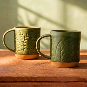 May include: Two green ceramic mugs with leaf and branch patterns. The mugs have brown bases and curved handles. The mugs are on a brown surface with a green background.