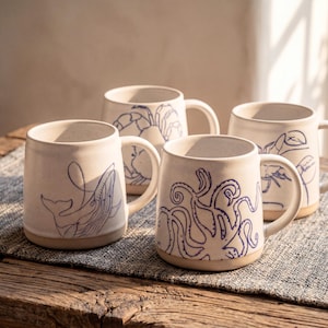 May include: Four cream-coloured ceramic mugs, each with a dark blue illustration of marine life: a whale, crab, lobster, and octopus. The mugs have handles and are displayed on a wooden surface. The mugs are suitable for hot drinks.