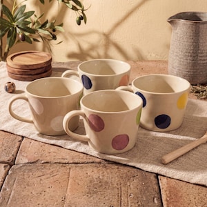Puede incluir: Cuatro tazas de cerámica beige con lunares grandes y coloridos. Cada taza tiene un asa y un punto de diferente color: rosa, azul, verde y amarillo. Una cuchara de madera y una jarra están en el fondo.
