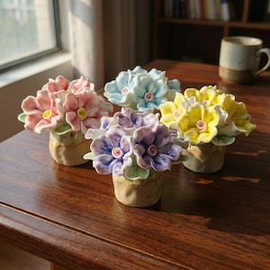 Könnte beinhalten: Eine Sammlung keramischer Blumenarrangements in verschiedenen Farben, darunter Rosa, Blau, Lila und Gelb. Jedes Arrangement zeigt detaillierte Blütenblätter und grüne Blätter, die in kleinen, strukturierten Töpfen angeordnet sind. Die Blumen stehen auf einer dunklen Holzoberfläche.