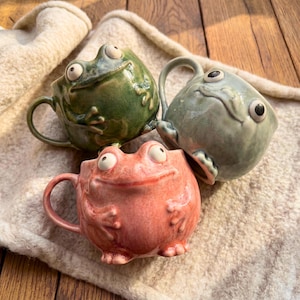 Puede incluir: Tres tazas de cerámica con forma de rana en tonos verdes, grises y rosas. Cada taza tiene un asa y ojos grandes y expresivos. Las tazas están sobre una superficie texturizada de color crema.