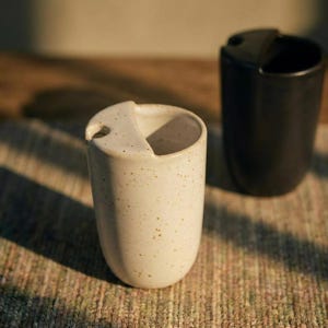 Może przedstawiać: Dwa ceramiczne kubki. Jeden jest w kolorze złamanej bieli z brązowymi plamkami i unikalnym kształtem uchwytu. Drugi kubek jest czarny z podobnym kształtem uchwytu. Oba kubki mają nowoczesną, minimalistyczną estetykę.