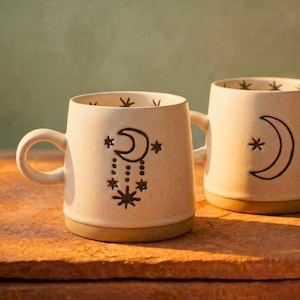 May include: Two cream ceramic mugs with a celestial design. One mug features a crescent moon and stars, the other has a similar design with a hanging moon and stars. Both mugs have a brown base and handle.