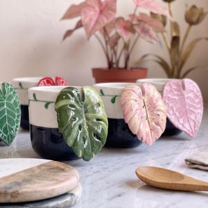 May include: A collection of ceramic mugs featuring a two-tone design with a dark blue base and a white upper section. Each mug is adorned with a three-dimensional, sculpted leaf handle in various colors, including green, pink, and red.