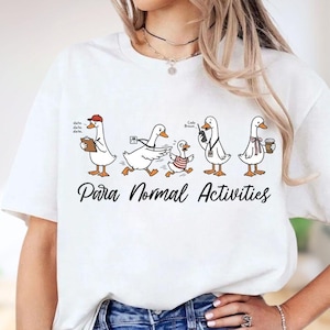 May include: A white t-shirt featuring a graphic of five cartoon ducks engaged in various activities with the text "Para Normal Activities" printed below. The ducks are depicted with accessories including a red cap, a clipboard, a radio, and a coffee cup.