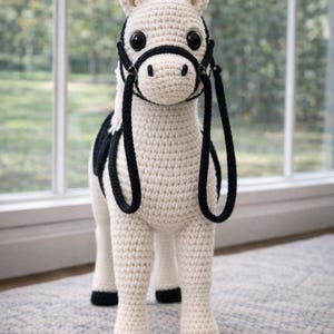 May include: A cream and black crochet horse toy. The horse has a black bridle and saddle, with black hooves. It stands on a light grey rug in front of a window. The toy is a soft, handmade item. The horse is a cute toy.