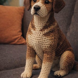 May include: A handcrafted crochet dog figurine in shades of brown and cream, sitting on a gray sofa. The dog has a detailed, textured appearance, with dark brown ears and a black nose. The background includes an orange pillow.
