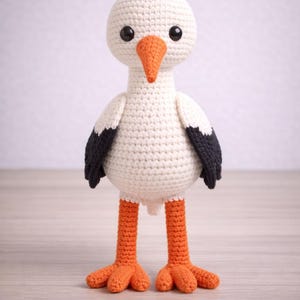 Puede incluir: Una muñeca de cigüeña de ganchillo hecha a mano. El pájaro tiene un cuerpo, cabeza y alas blancos con puntas de alas negras. Presenta un pico y patas naranjas y ojos negros. La muñeca está sobre una superficie de madera clara.