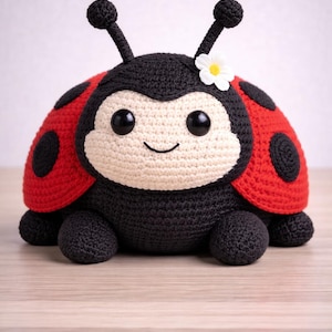 Puede incluir: Un juguete de crochet hecho a mano con forma de mariquita. La mariquita tiene un cuerpo negro, alas rojas con lunares negros y una cara color crema. Tiene ojos negros grandes, una boca sonriente y una pequeña flor blanca en la cabeza.