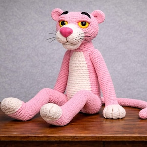 Może przedstawiać: Ręcznie robiona różowa lalka pantery z szydełka z białym brzuchem i łapami. Pantera ma żółte oczy, różowy nos i czarne wąsy. Siedzi na drewnianej powierzchni na szarym tle.