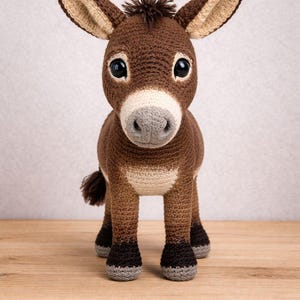 May include: A handmade crochet donkey toy in shades of brown, tan, and gray. The donkey has large, expressive eyes, long ears, and a small tail. It stands on four legs, with gray hooves, and is posed on a wooden surface.