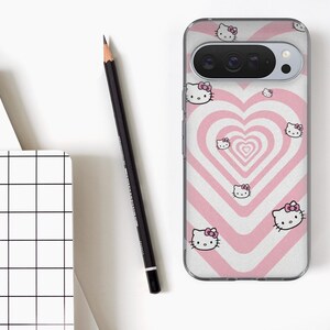 May include: A clear phone case featuring a pink and white heart design with multiple Hello Kitty character illustrations. A black pencil and a white notebook with a black grid pattern are also visible on a white surface.