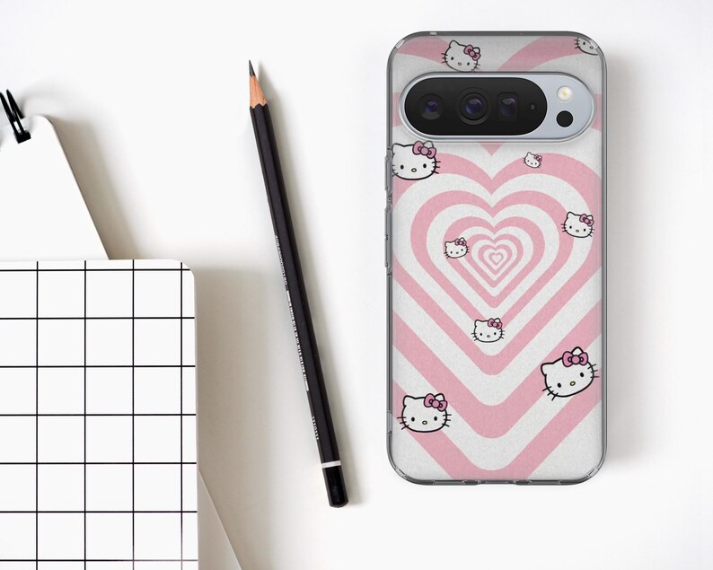 May include: A clear phone case featuring a pink and white heart design with multiple Hello Kitty character illustrations. A black pencil and a white notebook with a black grid pattern are also visible on a white surface.
