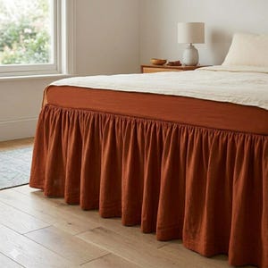 May include: A bed featuring a rust-coloured bed skirt and a cream-coloured bedspread. The bed skirt has a pleated design. The bed is in a room with a window and a lamp on a bedside table.