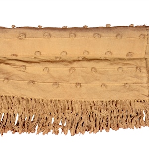 Può includere: Una coperta beige con un design strutturato e frange. La coperta presenta file orizzontali di piccoli cerchi in rilievo e una frangia lungo il bordo inferiore. Il colore è una tonalità calda e neutra.