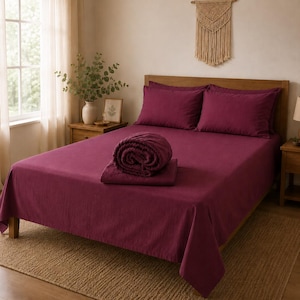 May include: A deep magenta bedding set featuring a flat sheet, two matching pillowcases, and a folded comforter placed in the center of the bed. The fabric has a visible woven texture, and the set is displayed on a wooden bed frame.