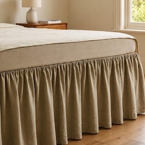Può includere: Una balza per letto color beige tinta unita con un design arricciato e a volant che si estende dalla base del materasso fino al pavimento. Il tessuto presenta un aspetto testurizzato e copre la parte inferiore della struttura del letto.