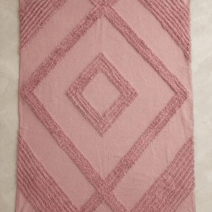 May include: A rectangular dusty rose throw blanket featuring a raised tufted diamond pattern across the surface. The blanket is finished with small decorative tassels at each of the four corners, providing a textured look to the fabric.