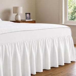 May include: A white bed skirt featuring a gathered, ruffled design that hangs from the base of the mattress to the floor. The fabric is solid white and opaque, providing a clean, uniform look that covers the lower frame of the bed completely.