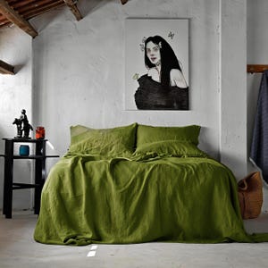 Puede incluir: Una cama con ropa de cama de lino verde oliva, que incluye una sábana bajera, una sábana encimera y fundas de almohada. La habitación tiene una pared blanca con una pintura de una mujer. Una mesita de noche negra contiene objetos decorativos.