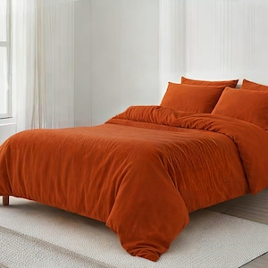 Puede incluir: Un juego de cama de color naranja, que incluye una funda nórdica y fundas de almohada, se muestra en una cama. La ropa de cama tiene un aspecto texturizado. La cama está en una habitación bien iluminada con una ventana y una pequeña mesita de noche de madera.