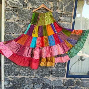 May include: A long, tiered skirt featuring a patchwork design of various colorful fabrics in shades of green, pink, red, orange, yellow, and blue. The skirt has an elasticized waistband and a flared, voluminous silhouette with multiple horizontal panels.