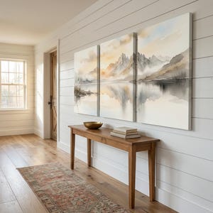 May include: A triptych canvas wall art set featuring a watercolor landscape of jagged mountains reflected in a calm lake. The scene uses soft tones of beige, gray, and pale blue, depicting a serene mountain range with misty clouds and water.