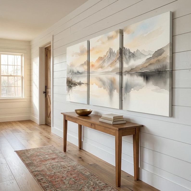 May include: A triptych canvas wall art set featuring a watercolor landscape of jagged mountains reflected in a calm lake. The scene uses soft tones of beige, gray, and pale blue, depicting a serene mountain range with misty clouds and water.