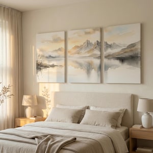 May include: A triptych wall art set featuring a watercolor landscape of misty mountains reflected in a calm lake. The artwork uses a soft palette of beige, grey, and muted gold tones, depicting jagged peaks, low clouds, and shoreline reeds.