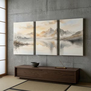 May include: A triptych wall art set featuring a watercolor landscape of jagged mountains reflected in a calm lake. The scene uses a soft palette of beige, grey, and white tones, depicting misty peaks, shoreline reeds, and a serene, hazy sky.