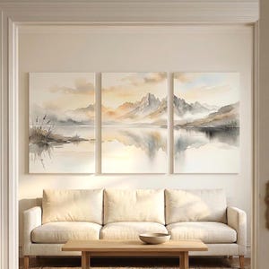 May include: A triptych wall art set featuring a watercolor landscape of jagged mountains reflected in a calm lake. The color palette consists of soft beige, muted gray, and pale blue tones, depicting a serene mountain range with misty clouds.