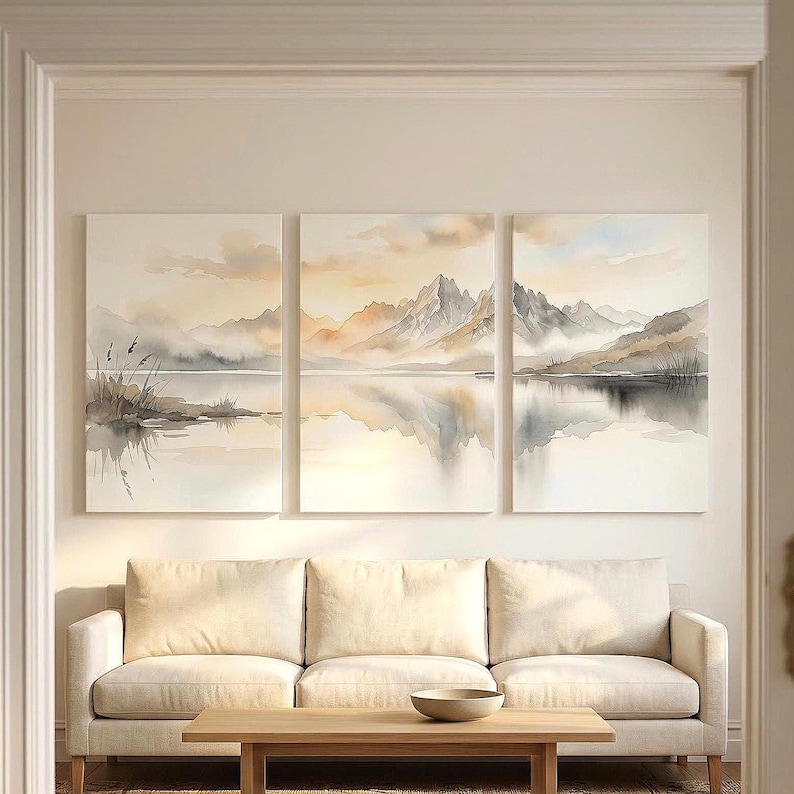 May include: A triptych wall art set featuring a watercolor landscape of jagged mountains reflected in a calm lake. The color palette consists of soft beige, muted gray, and pale blue tones, depicting a serene mountain range with misty clouds.