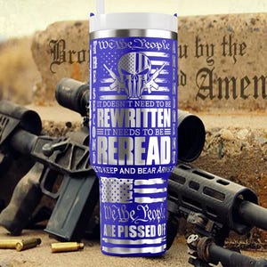May include: A blue and silver travel mug with the text "We the People" and "It doesn't need to be rewritten, it needs to be reread." The mug features a skull and crossbones design and American flag imagery. Firearms and ammunition are in the background.