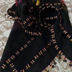 Peut inclure: Écharpe noire avec un motif floral rose, jaune et violet. L'écharpe a une bordure décorative avec de petits détails brodés colorés. L'écharpe est faite d'un tissu léger et est probablement portée comme accessoire de mode.