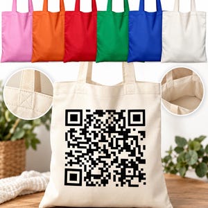 May include: A collection of canvas tote bags in pink, orange, red, green, blue, and off-white. The central off-white bag features a large black QR code printed on the front. Two circular insets show the reinforced handle stitching and interior.
