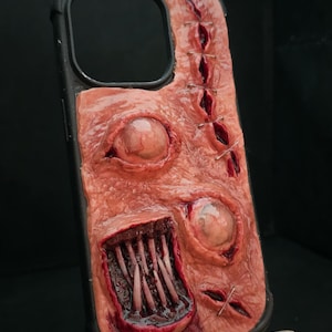 May include: A black phone case featuring a textured, flesh-toned surface with a realistic design. It includes two glossy, protruding eyeballs, a rectangular mouth with sharp, pointed teeth, and several stitched lacerations with metallic staples.