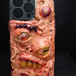May include: A black smartphone case featuring a textured, flesh-toned surface with multiple embedded realistic eyes and a set of yellowed teeth. The design includes deep red accents and organic, skin-like folds covering the back of the device.