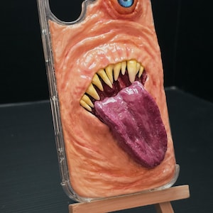 May include: A clear phone case featuring a three-dimensional, flesh-toned monster design with a single blue eye, a wide mouth filled with sharp yellow teeth, and a large, protruding purple tongue. The textured surface mimics skin folds and wrinkles.