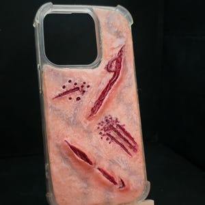 May include: A clear phone case featuring a textured, skin-toned surface with realistic-looking simulated wounds, including deep red lacerations, puncture marks, and scratch patterns, designed to mimic human flesh with a glossy, lifelike finish.