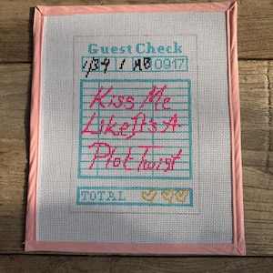 May include: A framed needlepoint piece with a pink border. The design features the words "Kiss Me Like It's A Plot Twist" in pink, set against a grid pattern. The words "Guest Check" are at the top.