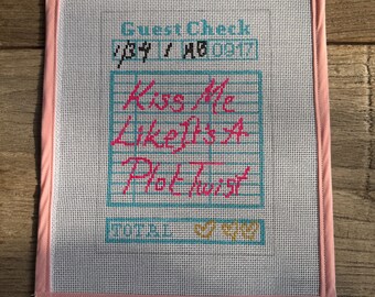 May include: A framed needlepoint piece with a pink border. The design features the words "Kiss Me Like It's A Plot Twist" in pink, set against a grid pattern. The words "Guest Check" are at the top.