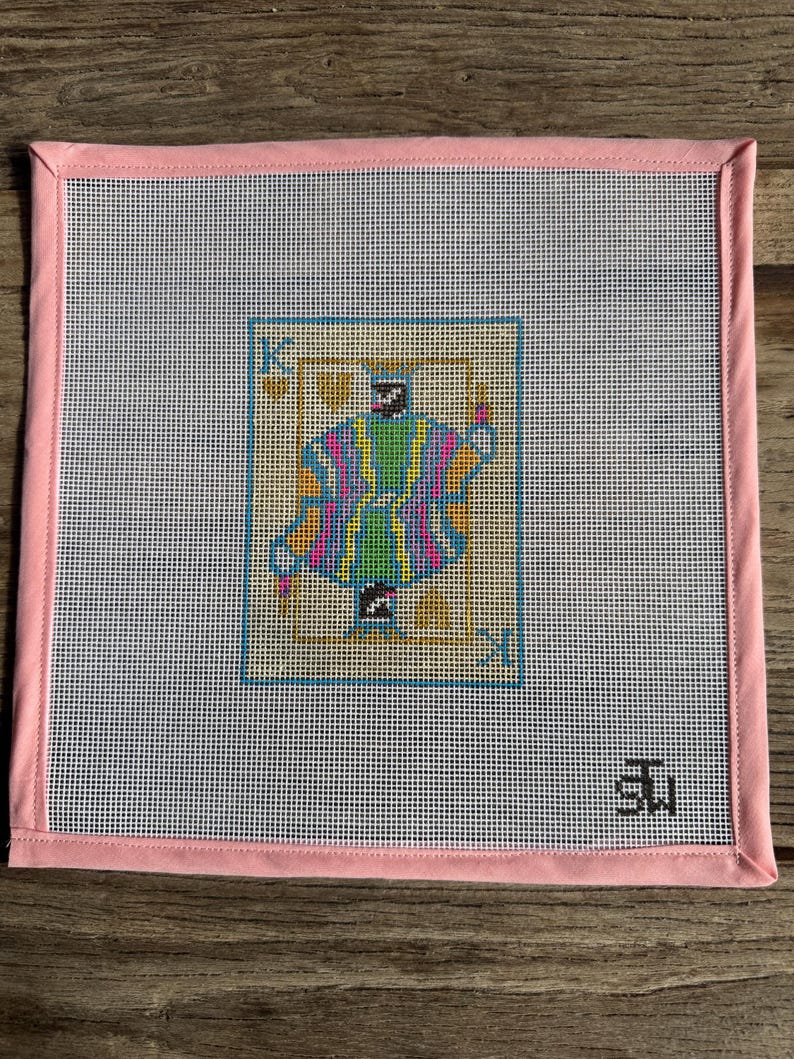 May include: Needlepoint canvas featuring a colorful King playing card design. The card has a blue border and is set against a white background. The canvas is framed with a pink border and is ready for stitching.