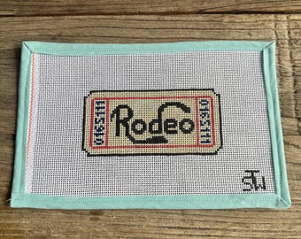 May include: A rectangular needlepoint canvas with a light blue border. The canvas features a ticket design with the word "Rodeo" in red and beige. The ticket has the numbers "0165111" and "0165111" on either side of the word "Rodeo".